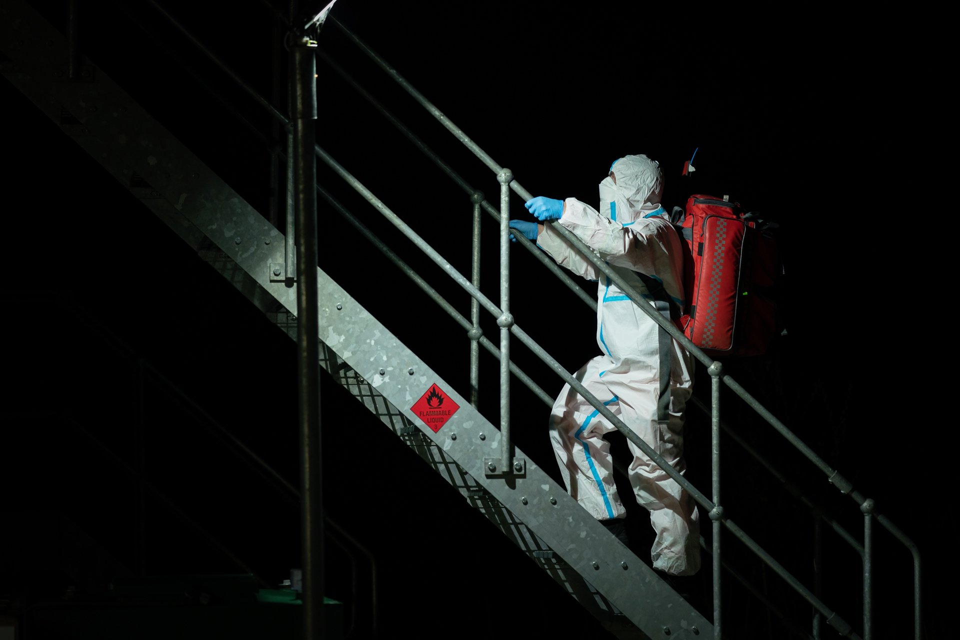 A photo of a paramedic in full hazmat PPE ascending metal stairs with a red kit bag on their back. A photo of a paramedic in full hazmat PPE ascending metal stairs with a red kit bag on their back.