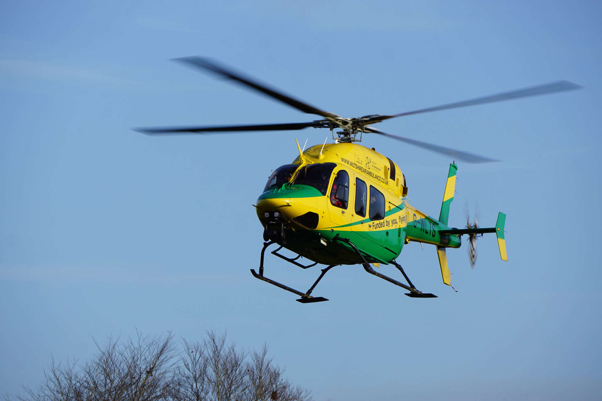 The yellow and green helicopter in flight against a blue sky with trees.