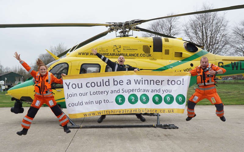 Two paramedics and a pilot jumping and smiling in front of the helicopter on a helipad. They are holding a large white and yellow banner advertising the Lottery Superdraw.