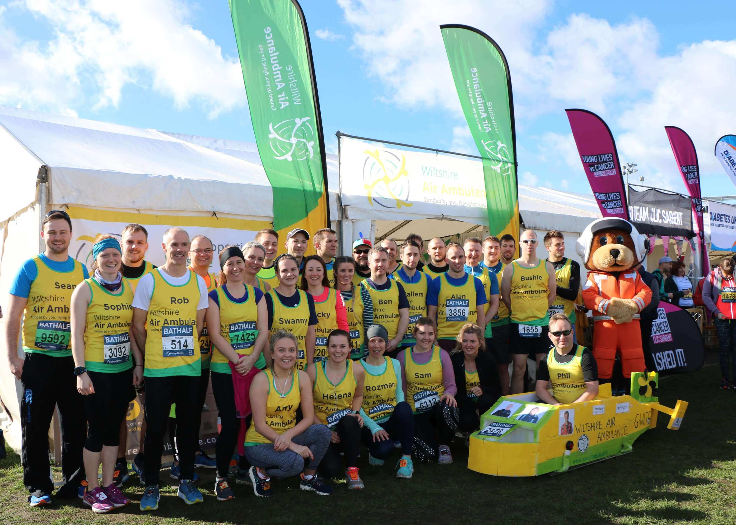 A group photo of participants together before running the Bath Half Marathon. All runners are wearing yellow and green running vests and are stood next to a large white gazebo.