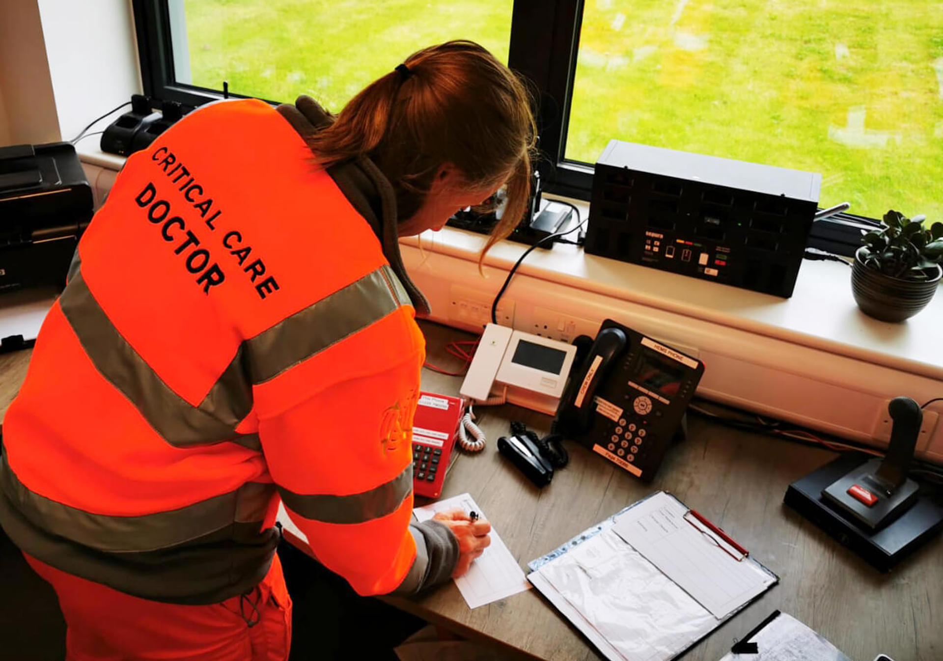 A doctor wearing an orange fleece taking notes whilst on a 999 call whilst stood at a wooden desk with paperwork and a clipboard.