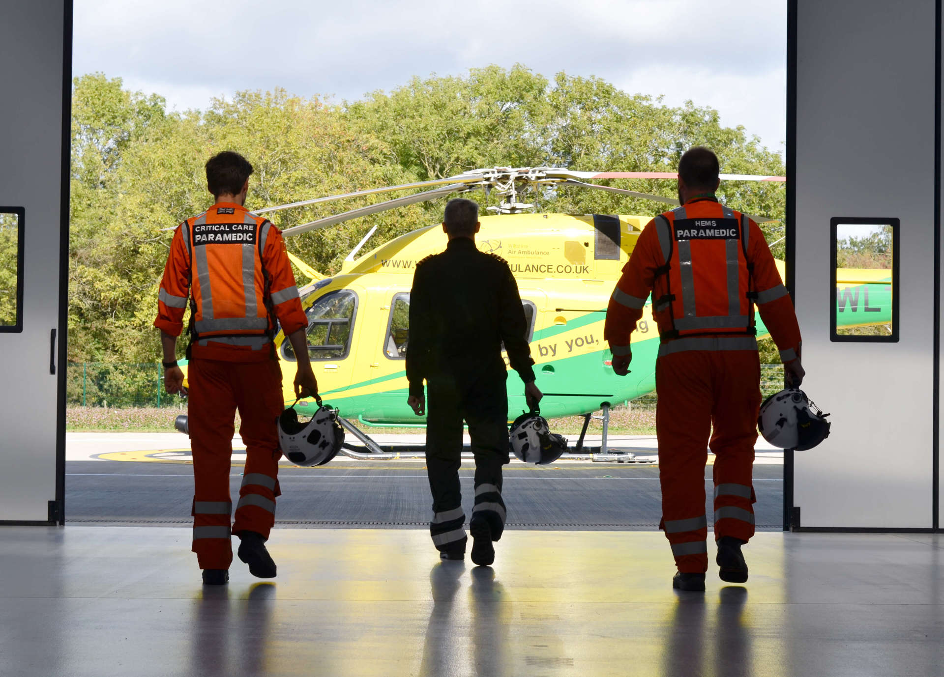 Two paramedics and a pilot walking towards the helicopter from inside the hangar.