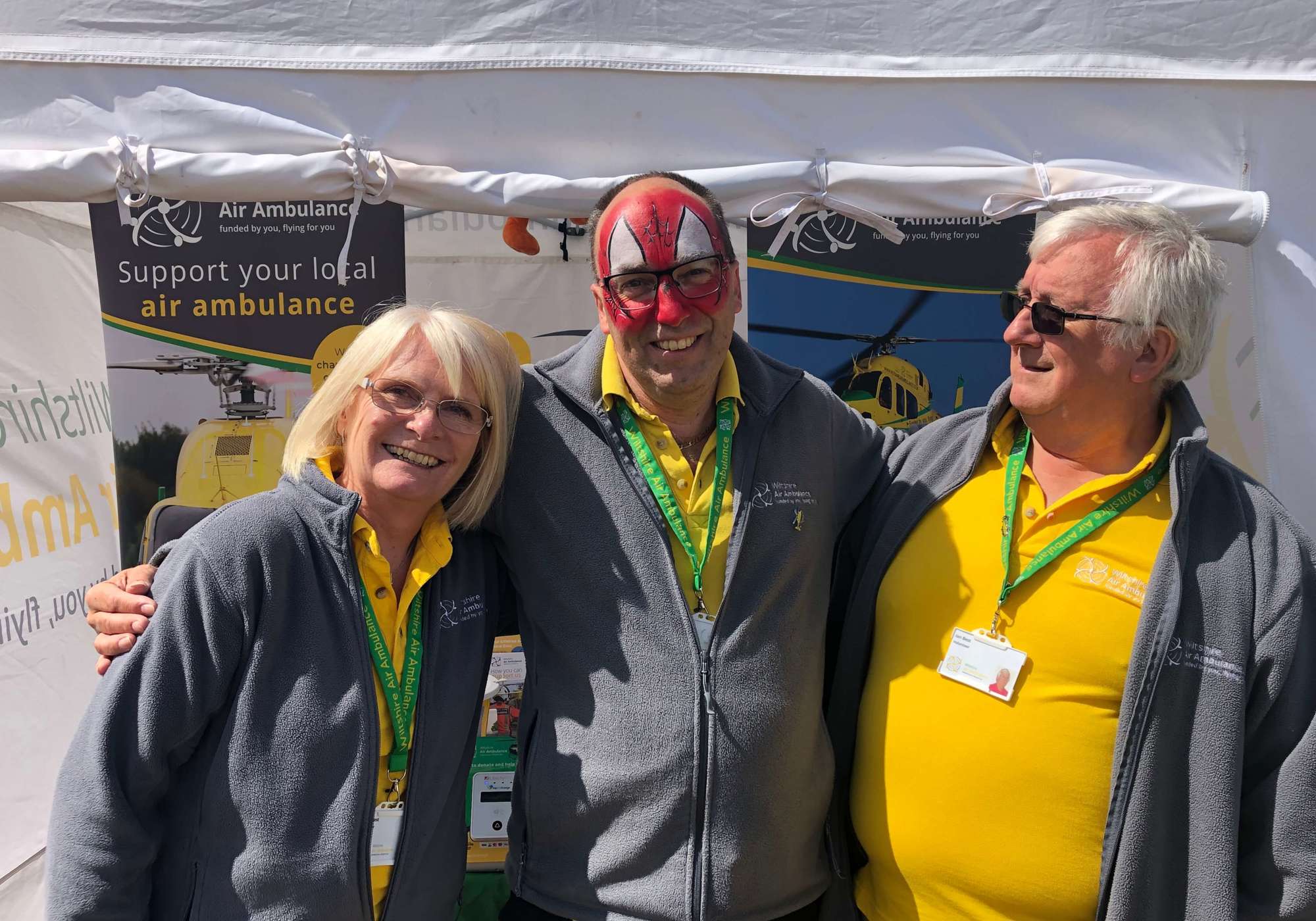 A group of three Wiltshire Air Ambulance volunteers wearing yellow polo shirts outside a gazebo at a fundraising event.