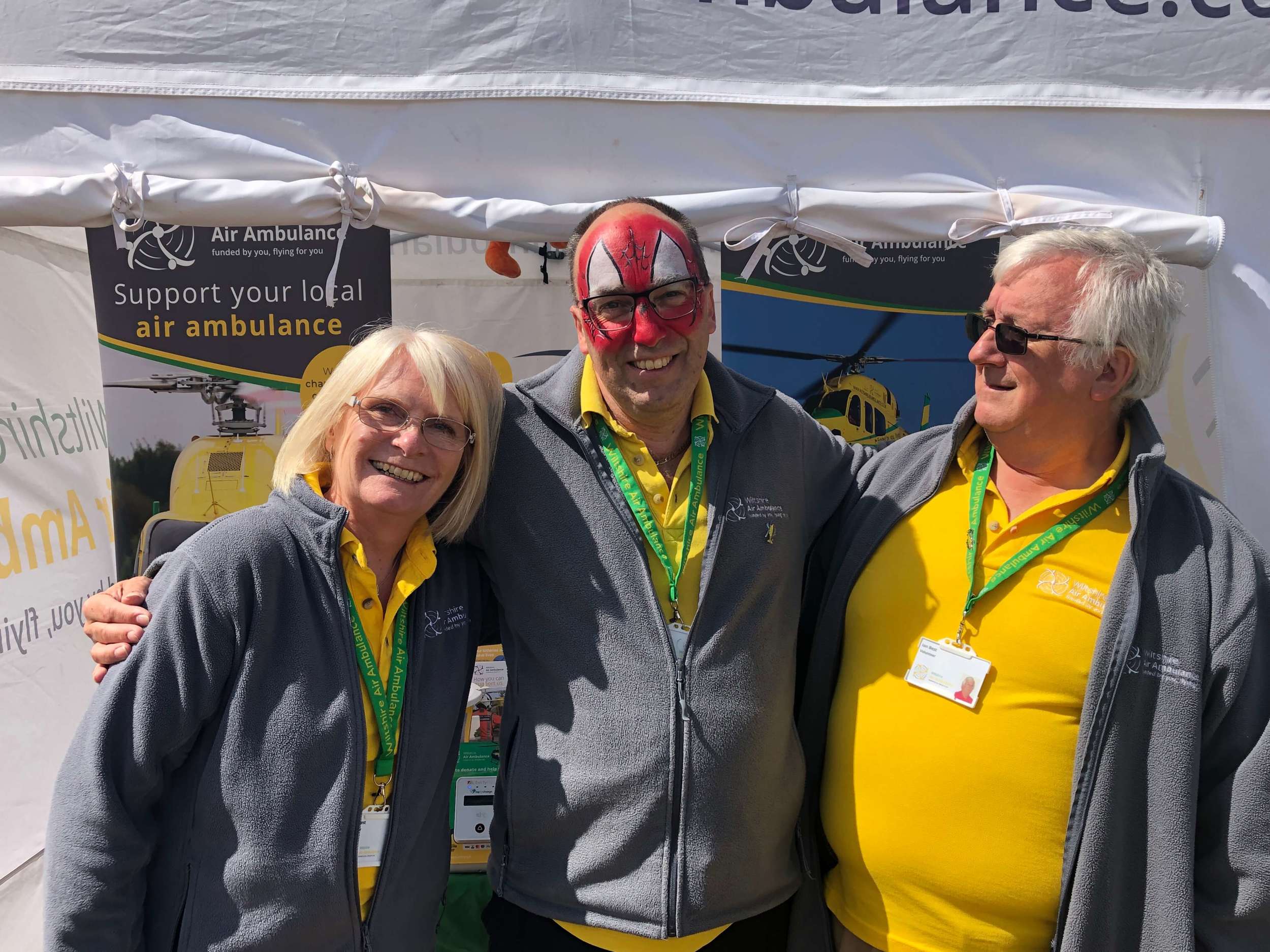 A group of three Wiltshire Air Ambulance volunteers wearing yellow polo shirts outside a gazebo at a fundraising event.