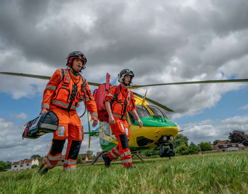 Two paramedics wearing orange flights walking away from the helicopter that has landed in a grass field. They are both carrying medical bags and equipment.