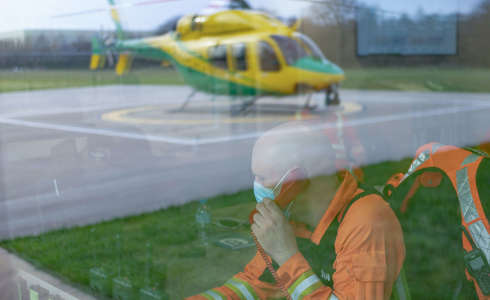 A paramedic wearing an orange flight suit answering a phone call in the flight room. There is a reflection of the helicopter on the helipad in the window.