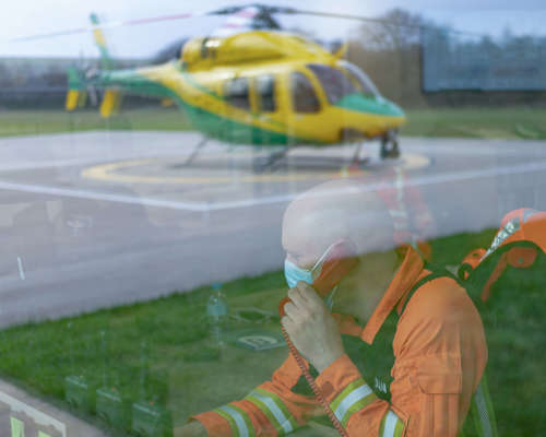 A paramedic wearing an orange flight suit answering a phone call in the flight room. There is a reflection of the helicopter on the helipad in the window.