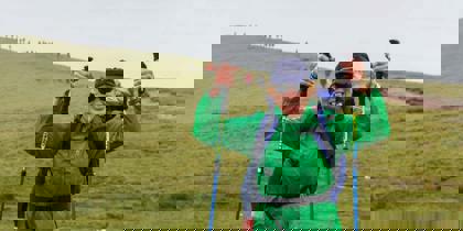 A person wearing a green raincoat and blue backpack, holding walking poles in the air