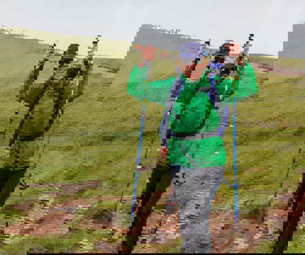 A person wearing a green raincoat and blue backpack, holding walking poles in the air