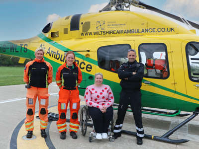 Two paramedics wearing orange flight suits and a pilot wearing a black flight suit stood with new ambassador Louise Hunt in front of the helicopter on a sunny day. Two paramedics wearing orange flight suits and a pilot wearing a black flight suit stood with new ambassador Louise Hunt in front of the helicopter on a sunny day.
