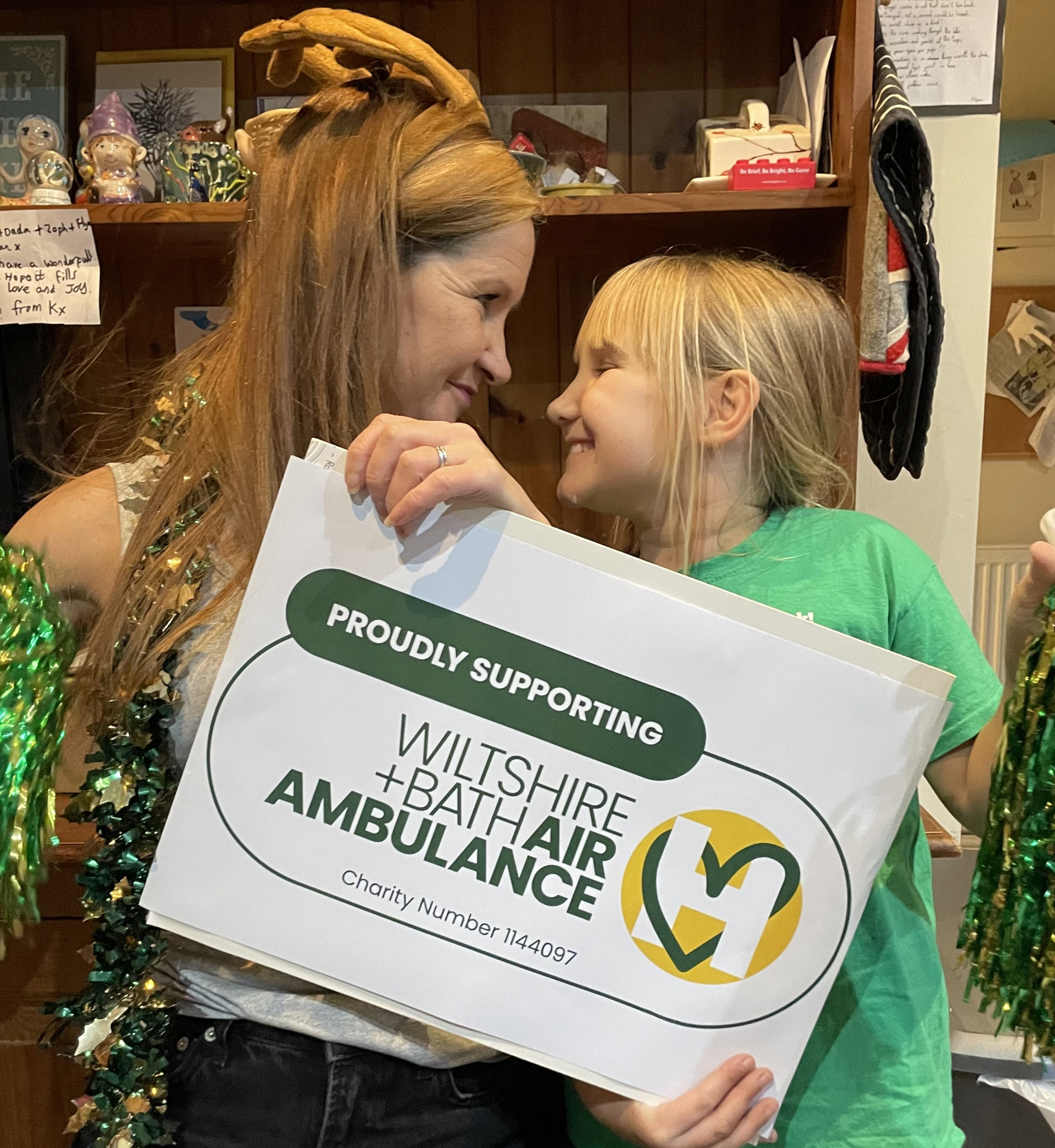 A lady and girl holding a proudly supporting Wiltshire and Bath Air Ambulance logo, whilst wearing greeb A lady and girl holding a proudly supporting Wiltshire and Bath Air Ambulance logo, whilst wearing greeb