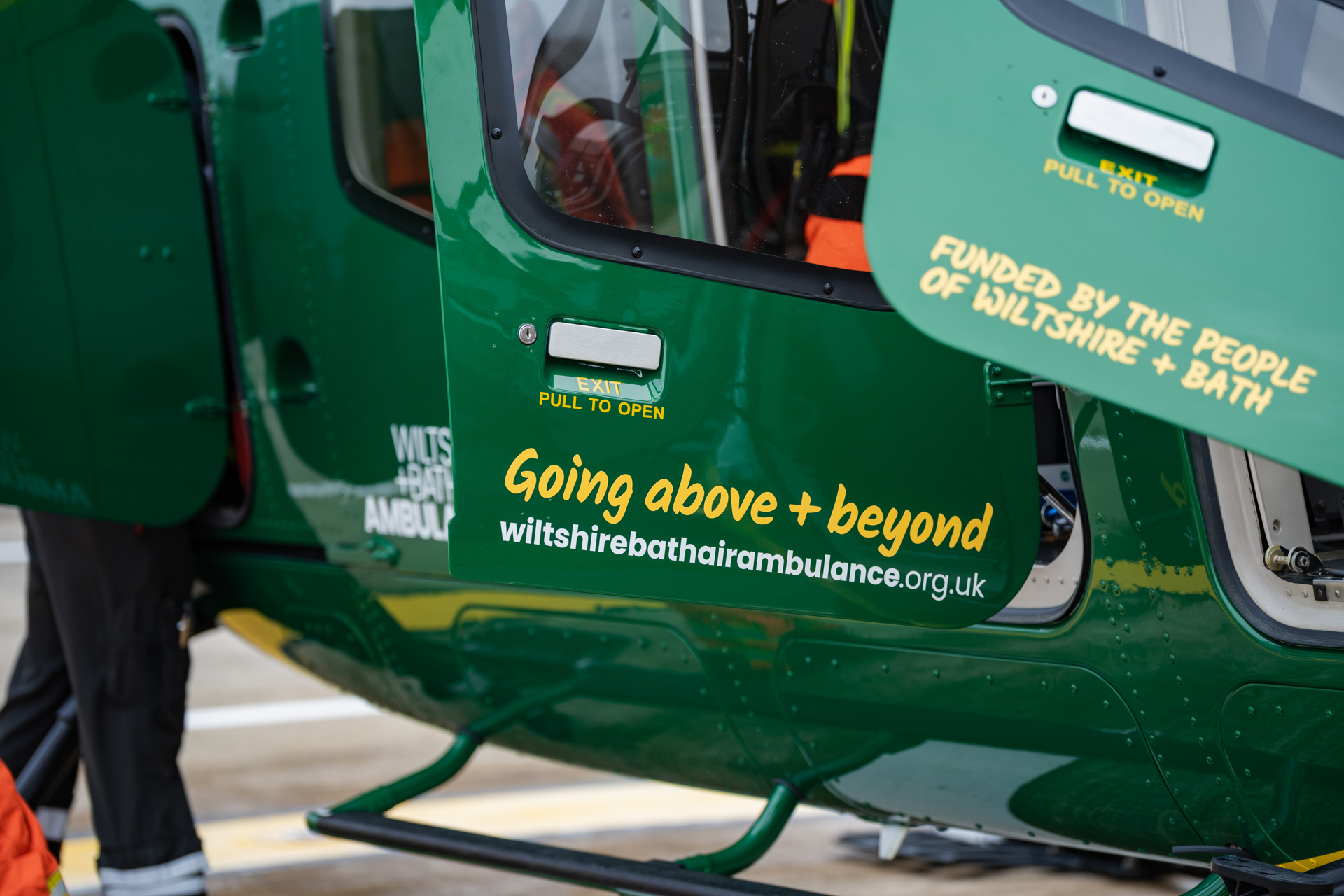 Yellow text with going above + beyond and funded by the people of wiltshire + bath on the doors of a green helicopter Yellow text with going above + beyond and funded by the people of wiltshire + bath on the doors of a green helicopter