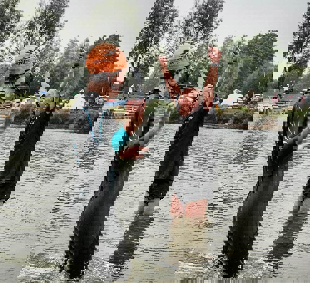 A swimmer wearing a wet suit and orange swim hat, standing knee deep in water, with a man behind wearing a t-shirt and shorts with his arms in the air cheering people on