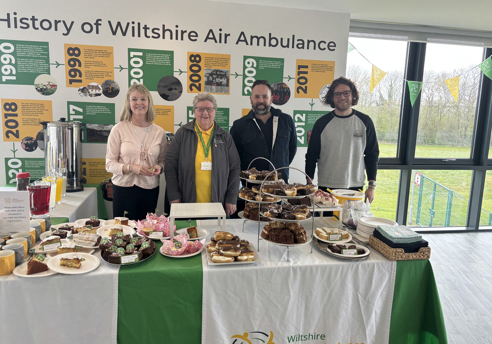 A table full of cakes, with four adults standing behind it, smiling at the camera