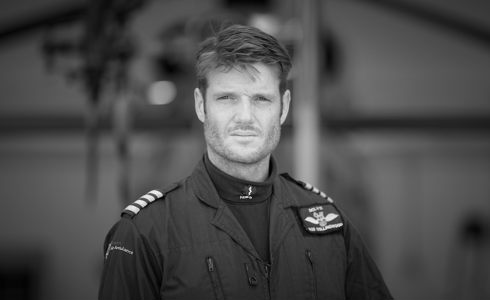 A black and white photograph of a pilot stood in front of the helicopter wearing a flight suit.