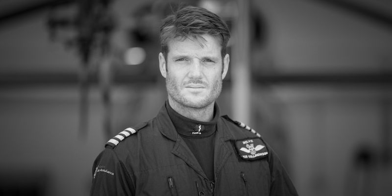 A black and white photograph of a pilot stood in front of the helicopter wearing a flight suit.