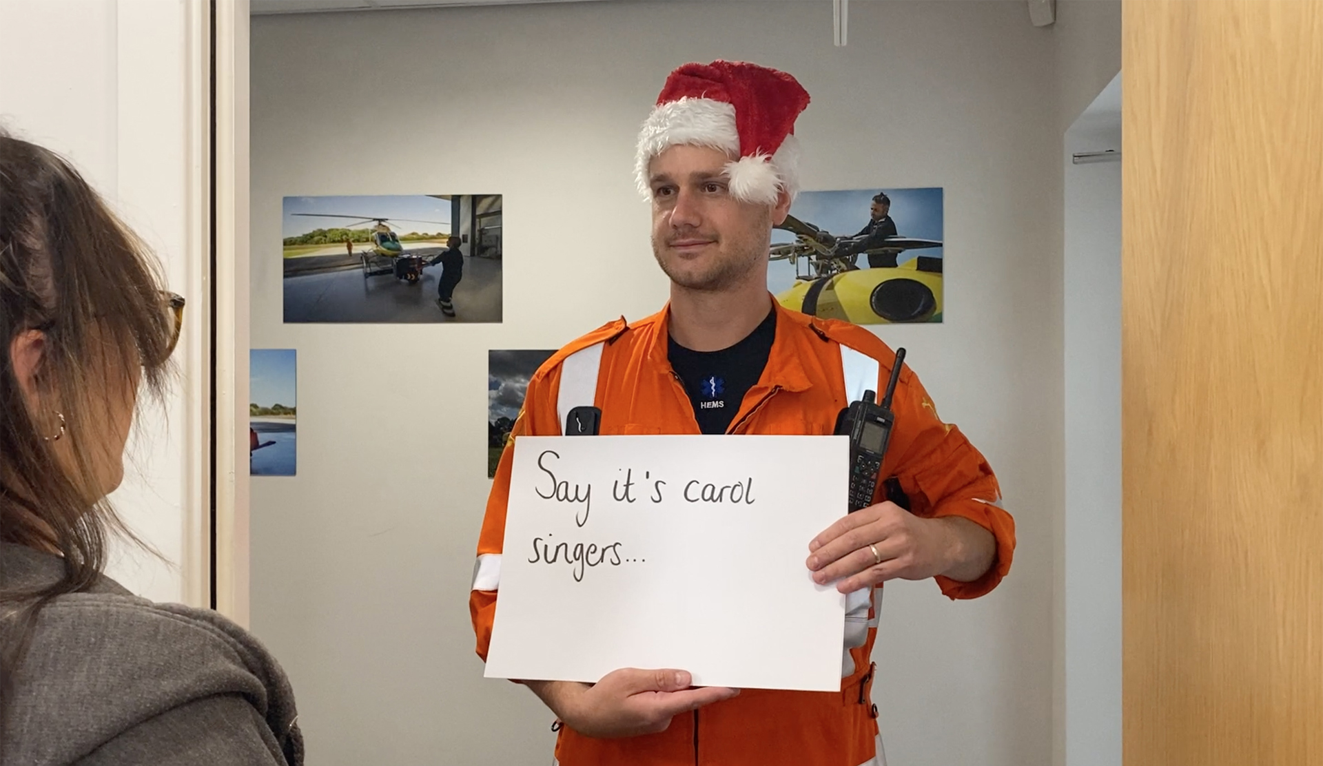 A paramedic wearing a santa hat, holding a "say it's carol singers" sign