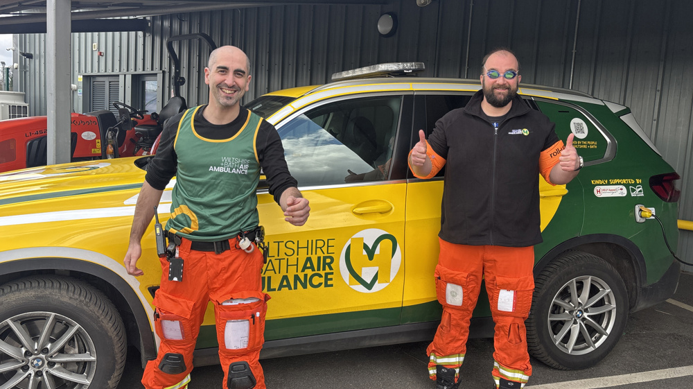 Two men standing in front of a yellow and green car, one wearing a running vest and the other wearing swimming goggles and armbands